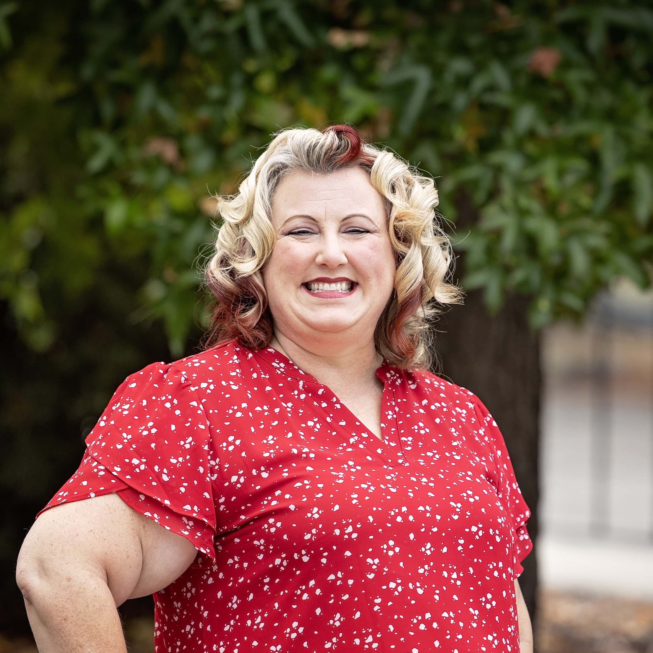 Woman with curled blonde and red hair smiling outdoors in front of leafy trees, wearing a bright red blouse with small white floral patterns.