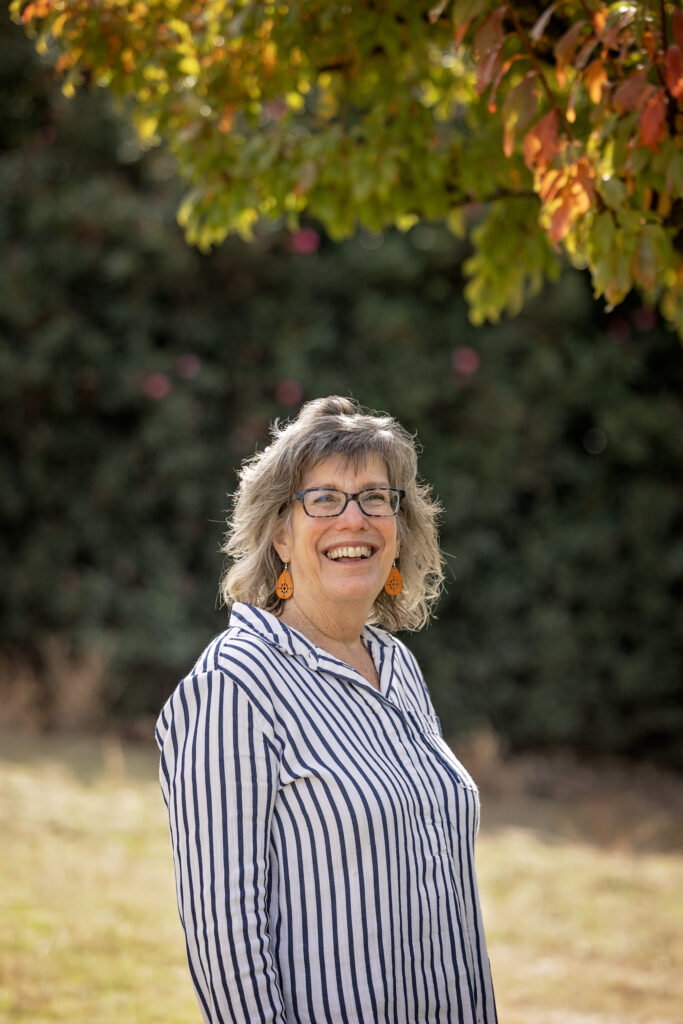A woman with gray hair and glasses smiles while standing under a tree with green and orange leaves. She is wearing orange earrings and a blue-and-white striped blouse.