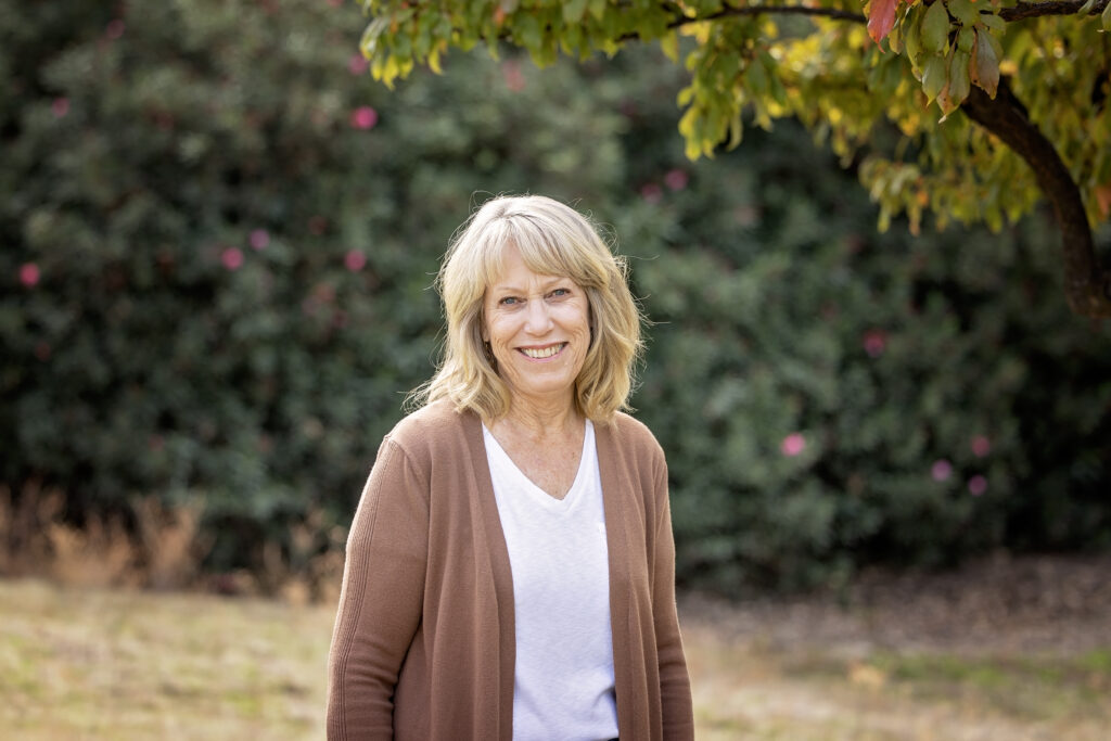 A woman with shoulder-length blonde hair smiles warmly outdoors beneath a tree with light green leaves. She is wearing a white shirt and a light brown cardigan.