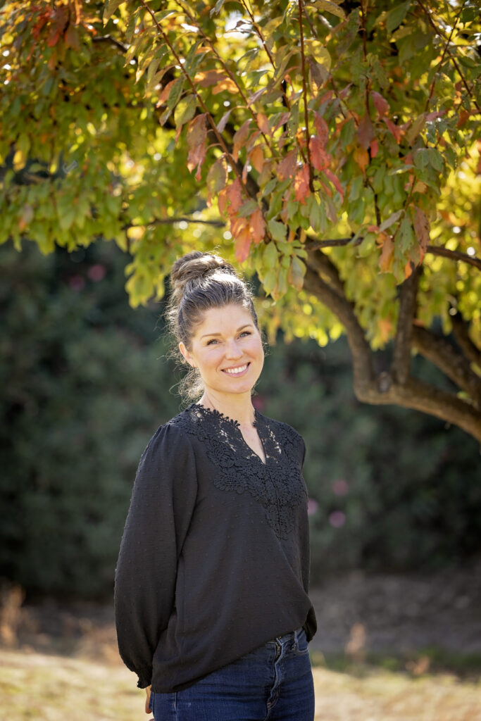 A woman with her brown hair in a bun smiles while standing beneath a tree with autumn-colored leaves. She is wearing a black long-sleeve blouse with lace detailing and blue jeans.