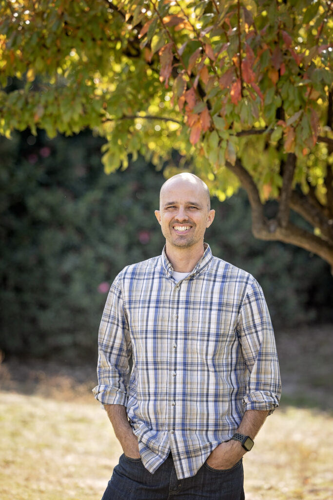 A man with a shaved head smiles while standing outdoors under a tree with green and orange leaves. He is wearing a beige and blue plaid button-up shirt.