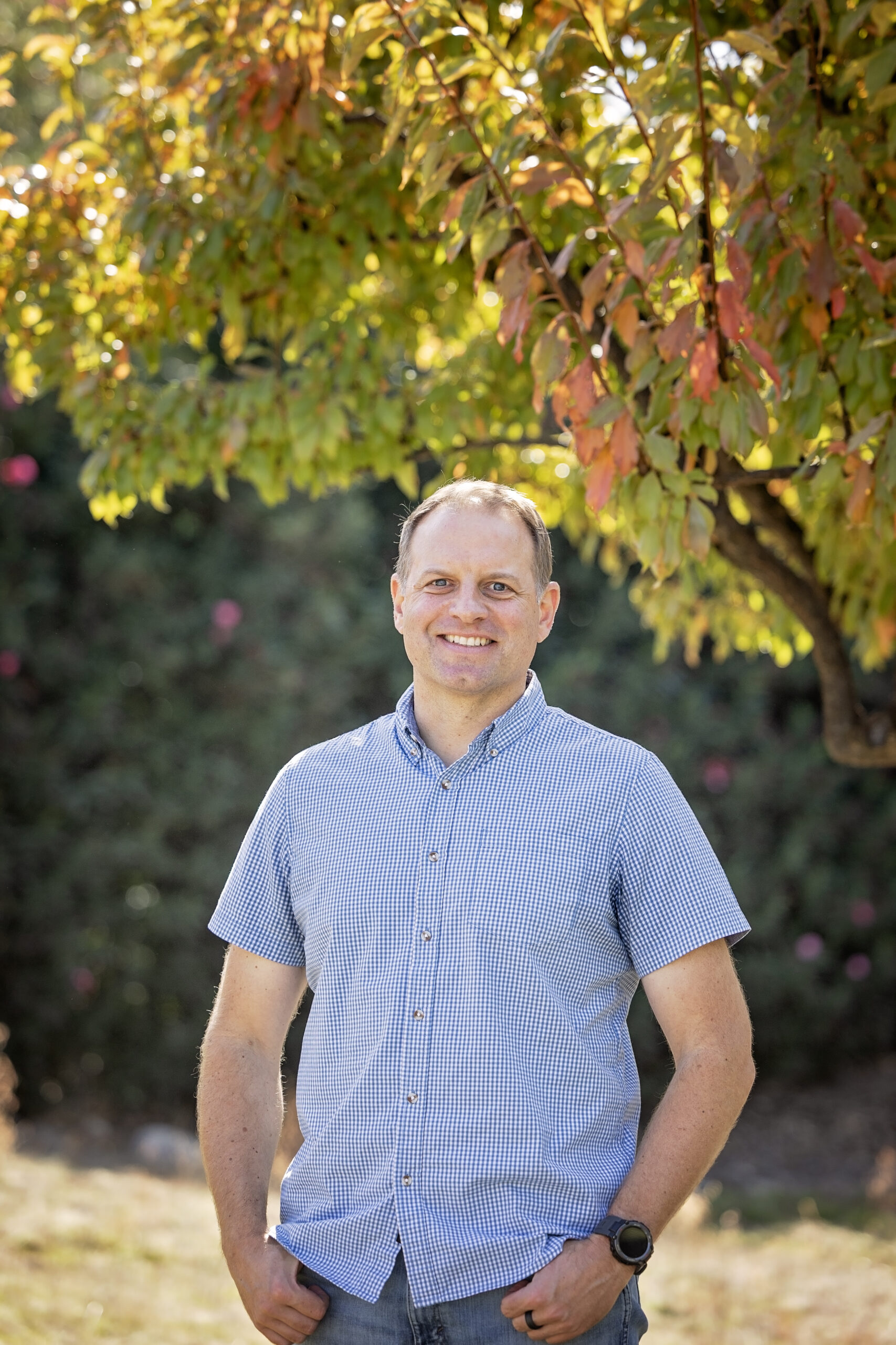 Man smiling outdoors beneath a tree with green and orange leaves, wearing a short-sleeve blue and white checkered button-up shirt and jeans.