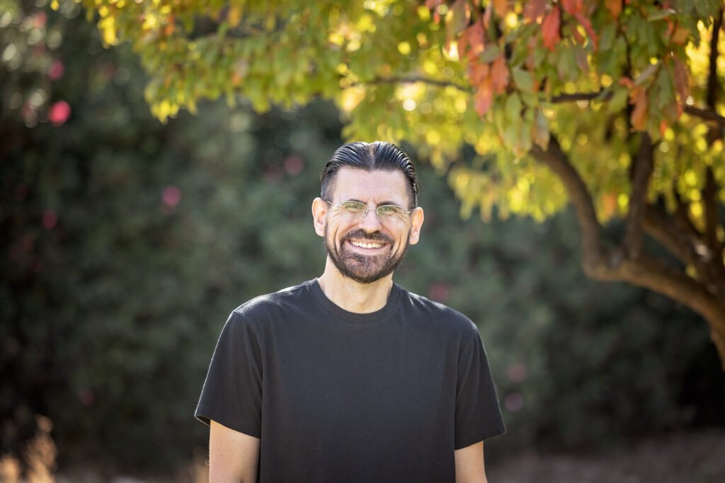 A man with dark hair and a beard smiles while standing beneath a tree with fall-colored leaves. He is wearing glasses and a plain black T-shirt.