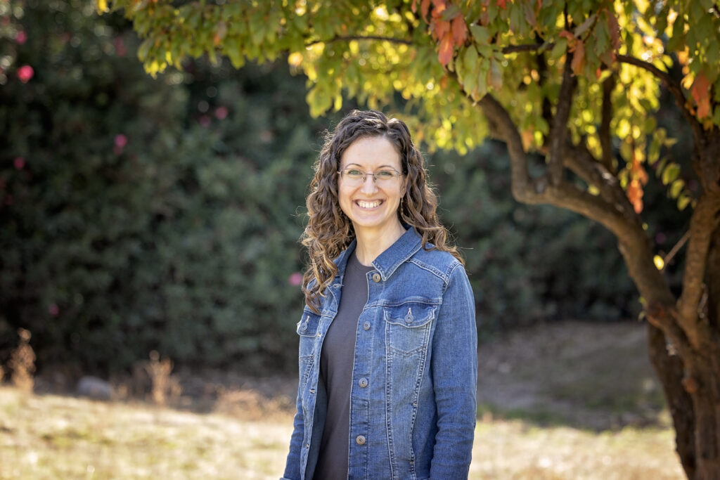 A woman with curly brown hair smiles outdoors under a tree with green and orange leaves. She is wearing glasses, a dark gray top, and a blue denim jacket.