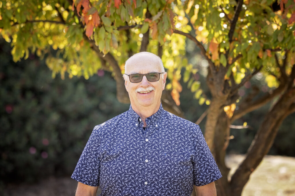 A man with a bald head, white mustache, and sunglasses smiles while standing beneath a tree with golden and orange leaves. He is wearing a navy blue short-sleeve shirt with a white abstract pattern.